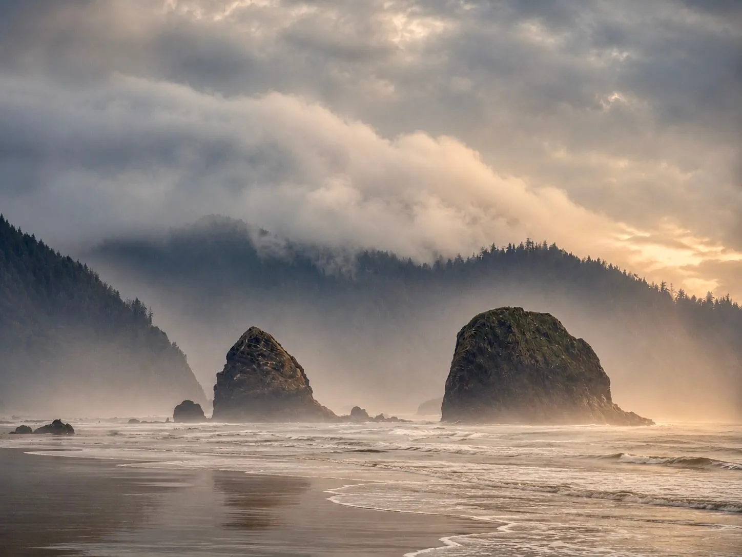 Two sea stacks in sunlit fog below a forested headland, surf running up the beach