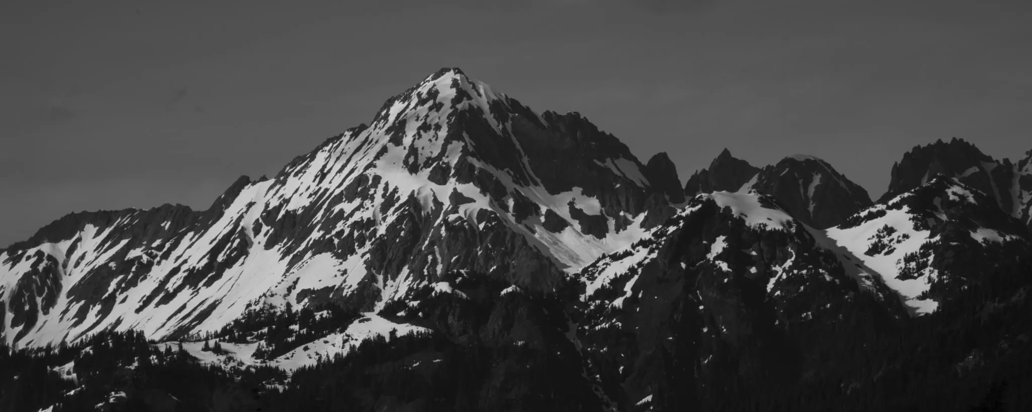 Black-and-white telephoto panorama of a sharp, snow-streaked peak and a jagged ridgeline above dark forest