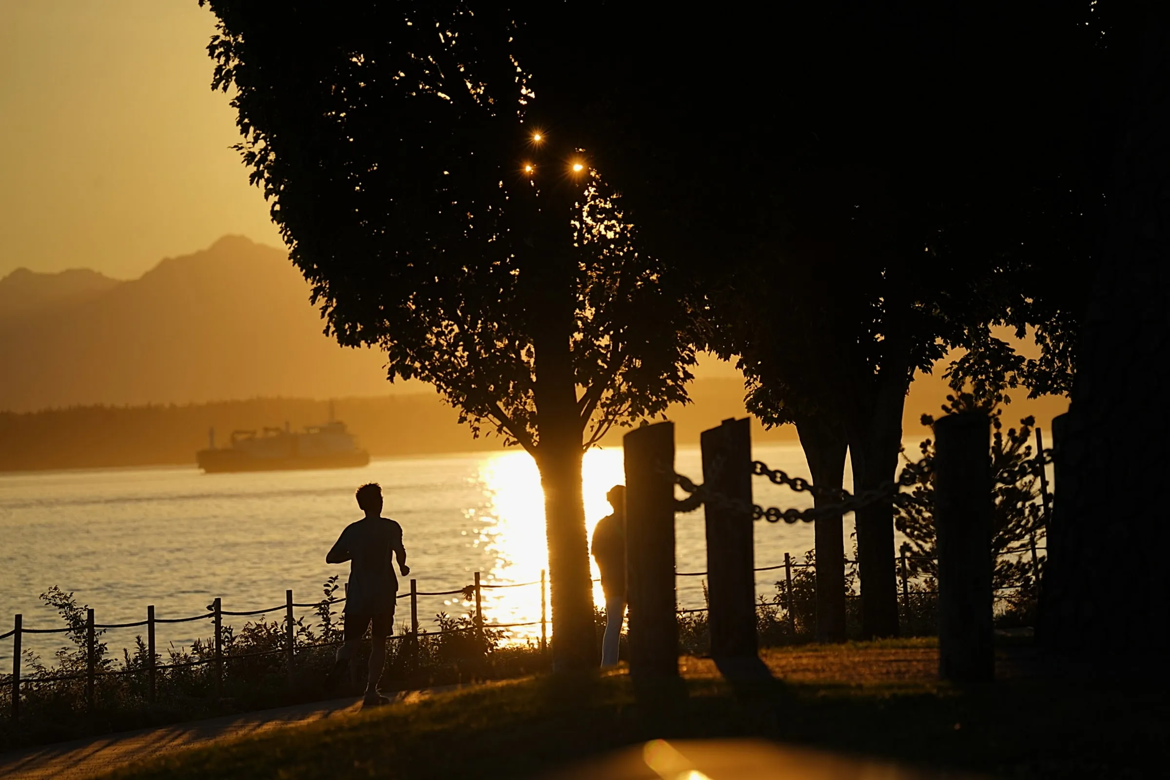 A runner silhouetted on a waterfront path at golden hour, a ferry and the Olympic Mountains across the water