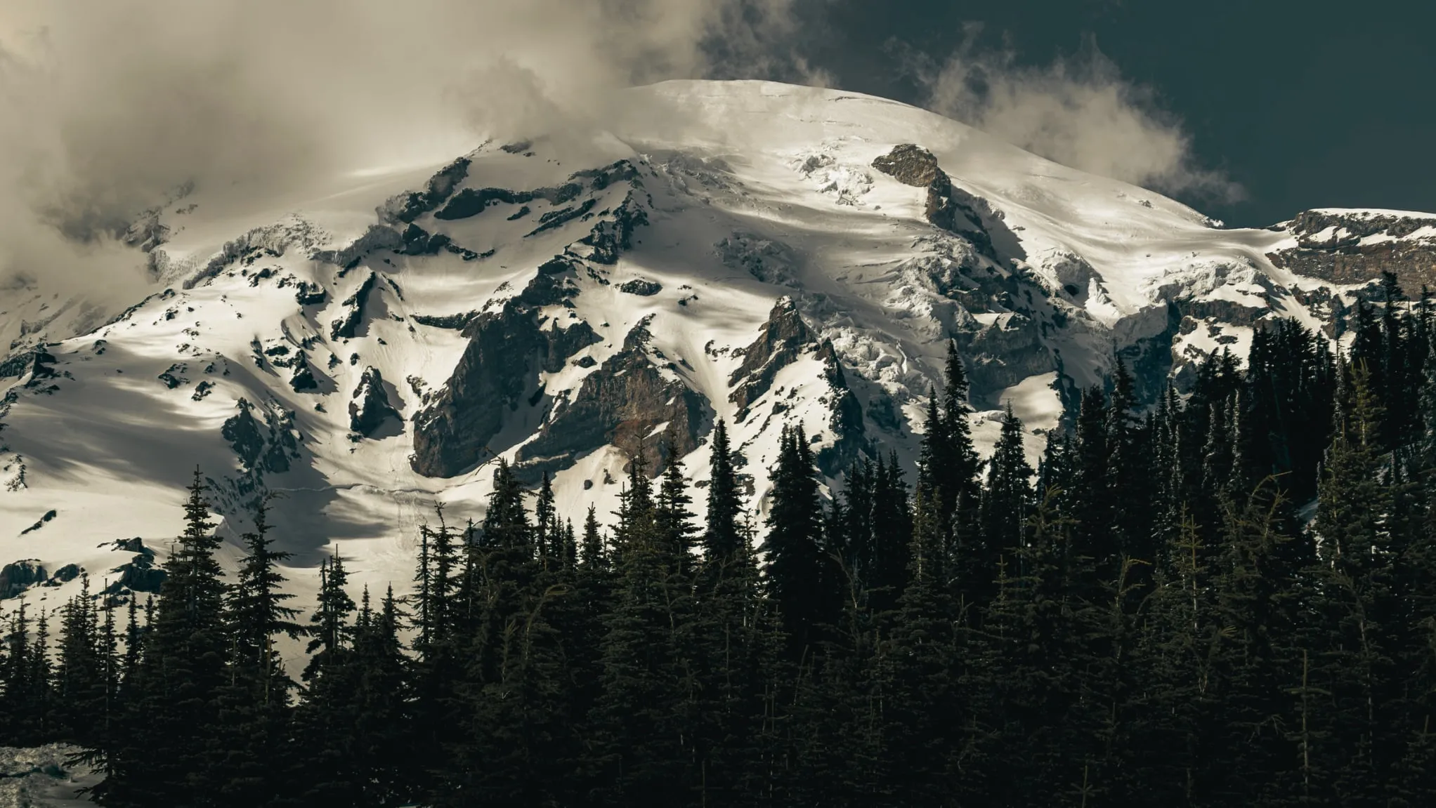 Close view of Mount Rainier’s glaciers and rock ribs under breaking cloud, dark firs below